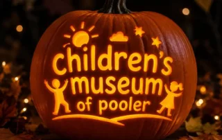 Glowing jack-o’-lantern carved with the Children’s Museum of Pooler logo, surrounded by autumn leaves and string lights