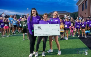 Three Heartwood team members in purple race shirts hold a $15,000 check made out to RHHS on the turf at Richmond Hill High School, with 5K runners cheering behind them
