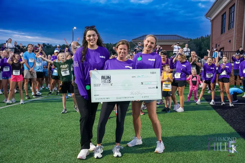 Heartwood Run for the Hill 5K 2026 check presentation Three Heartwood team members in purple race shirts hold a $15,000 check made out to RHHS on the turf at Richmond Hill High School, with 5K runners cheering behind them